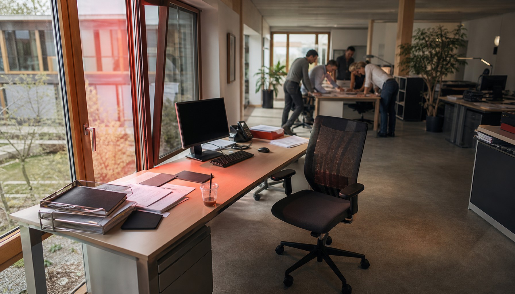 Leerer Bürostuhl vor einem Schreibtisch, im Hintergrund bespricht ein Team Unterlagen an einem Tisch.